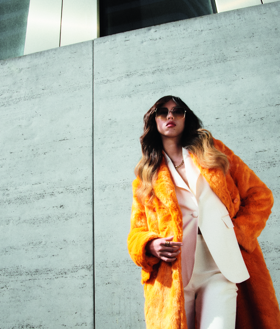 Mulher com casaco laranja e fato branco, de pé junto a uma parede de cimento