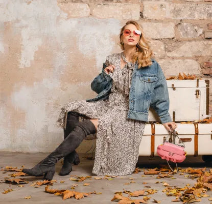 Mulher loira com look boho, a usar um vestido floral, casaca de ganga e botas, sentada sobre uma mala