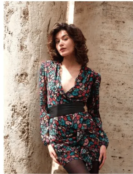 Uma mulher com cabelo escuro e ondulado, vestindo um vestido estampado com padrão floral em tons de azul, rosa e verde, com um cinto preto na cintura e meias pretas, posa encostada a uma parede de pedra texturizada. Ela olha para a câmara.