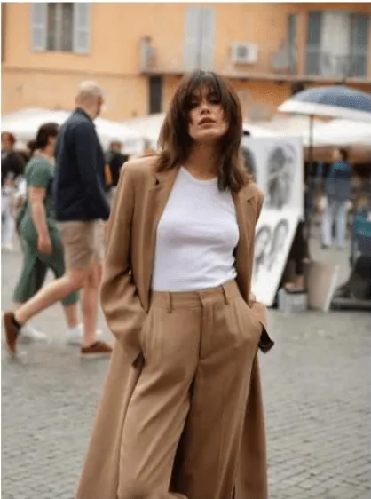 Uma mulher com cabelo castanho e franja, vestindo um casaco e calças de cor camel e uma t-shirt branca, posa com as mãos nos bolsos. Ela está de pé numa rua com pessoas e edifícios desfocados ao fundo.