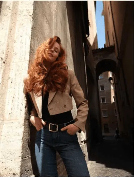 Uma mulher com cabelo ruivo longo e ondulado, vestindo um top preto, um casaco cropped de padrão xadrez e calças de ganga com um cinto, posa num beco estreito entre edifícios de pedra. Ela tem as mãos nos bolsos e a cabeça ligeiramente inclinada para trás.