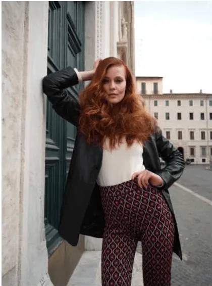 Uma mulher com cabelo ruivo longo e volumoso, vestindo um casaco de couro preto, uma camisola branca e calças com um padrão geométrico em tons de vermelho e castanho, posa encostada a uma porta verde. Ela tem uma mão na cabeça e a outra na anca, olhando para a câmara. Edifícios e um céu claro são visíveis ao fundo.