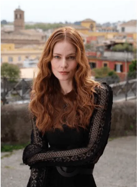 Uma mulher com cabelo ruivo longo e ondulado, vestindo uma blusa preta com detalhes em renda nas mangas e um cinto preto, posa ao ar livre. Ela olha diretamente para a câmara, com uma cidade italiana e uma torre de sino visíveis ao fundo.