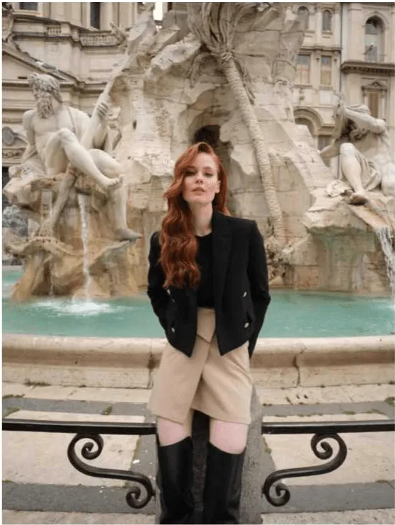 Uma mulher com cabelo ruivo longo e ondulado, vestindo um blazer preto, uma saia bege e botas pretas de cano alto, posa em frente a uma fonte esculpida com figuras mitológicas. Ela olha diretamente para a câmara.