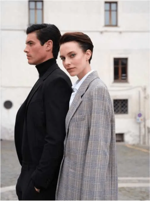 Um homem e uma mulher posam de costas um para o outro. O homem, de perfil, veste um casaco preto e uma camisola de gola alta preta. A mulher, com cabelo curto e escuro, veste um blazer cinzento de padrão xadrez e uma camisa branca, e olha para a câmara por cima do ombro.
