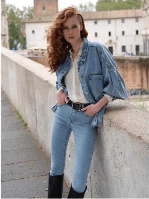Uma mulher ruiva com cabelo comprido e ondulado, vestindo um casaco de ganga, camisa branca, calças de ganga e botas pretas, posa numa ponte.