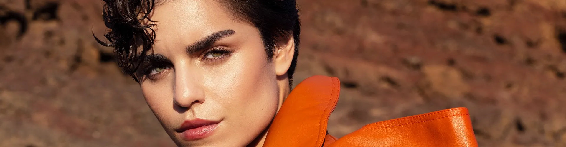 Um close-up do rosto de uma mulher com cabelo escuro e curto, sobrancelhas fortes e olhos verdes, vestindo uma gola de casaco cor de laranja.