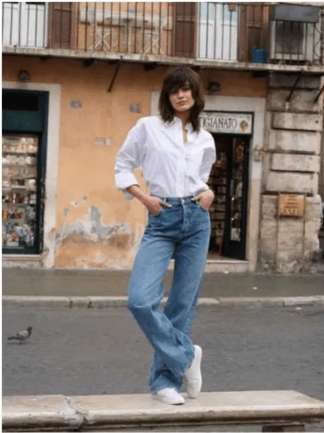 Uma mulher com cabelo castanho com franja, vestindo uma camisa branca e calças de ganga largas com ténis brancos, posa com as mãos nos bolsos. Ela está em pé numa estrutura baixa em frente a um edifício com paredes cor de pêssego e janelas escuras. Há um pombo no chão à esquerda.