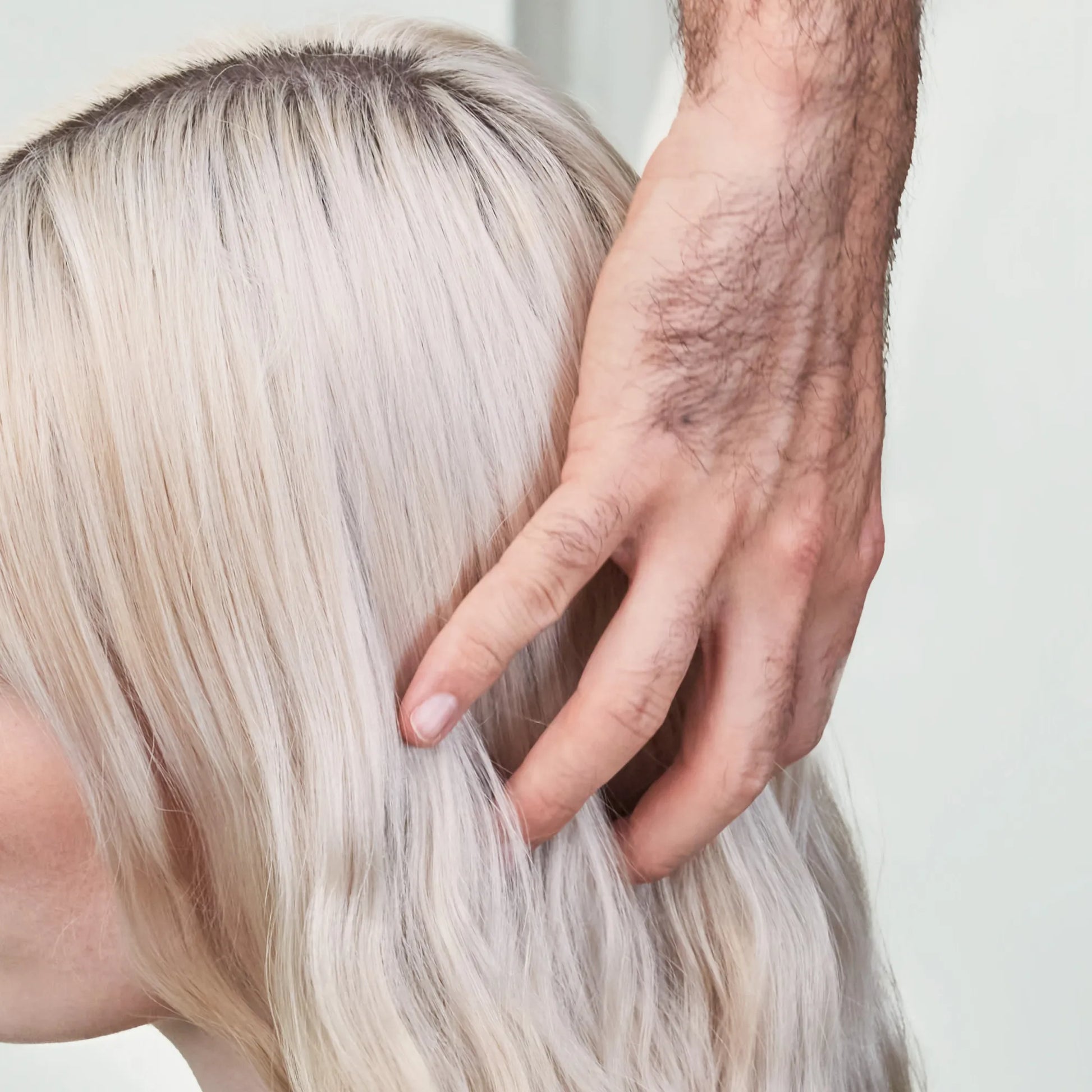 Uma mão masculina com pelos no braço a tocar gentilmente no cabelo louro ondulado de uma pessoa loira.