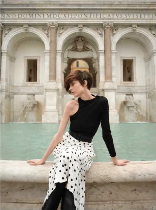 Uma mulher com cabelo curto castanho avermelhado, vestindo uma camisola preta de um ombro só e uma saia branca com bolinhas pretas, está sentada na beira de uma fonte. Atrás dela, há um edifício clássico com arcos e esculturas.