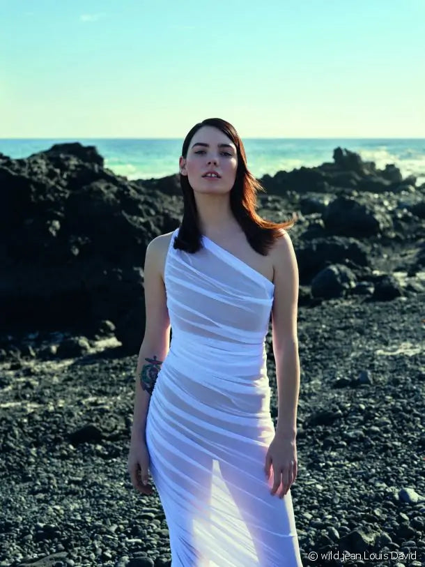 Uma mulher com cabelo escuro comprido e liso, vestindo um vestido branco transparente de um ombro só, posa numa praia rochosa com o oceano ao fundo.