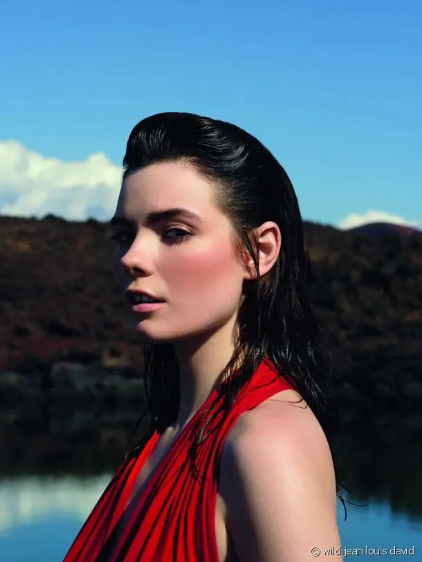 Um close-up de uma mulher com cabelo escuro e húmido penteado para trás, vestindo um top vermelho, com um lago e paisagem montanhosa ao fundo.