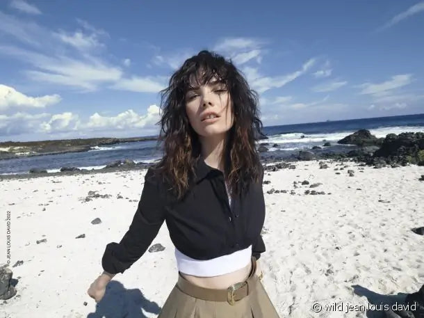 Uma mulher com cabelo escuro, vestindo uma camisa preta, um top branco e uma saia bege, posa numa praia arenosa com o oceano e o céu azul ao fundo.