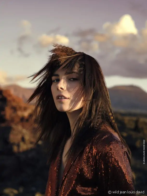 Uma mulher com cabelo castanho comprido e despenteado, vestindo uma blusa cor de cobre brilhante, posa ao ar livre com montanhas e céu nublado ao fundo.
