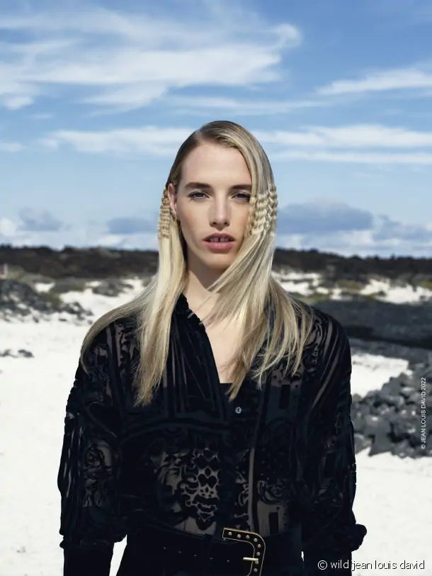 Uma mulher loira com cabelo liso, vestindo uma blusa preta transparente, posa numa paisagem árida com céu azul e algumas nuvens.