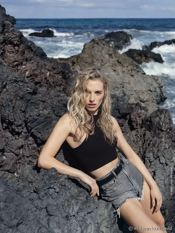 Uma mulher loira com cabelo ondulado, vestindo um top preto e calções de ganga, posa sentada em rochas à beira-mar.
