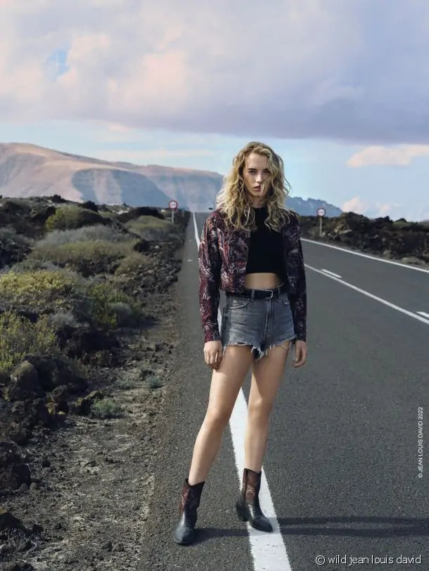 Uma mulher loira com cabelo encaracolado, vestindo um top preto, casaco estampado, calções de ganga e botas escuras, está de pé numa estrada deserta. Ao fundo, há montanhas e céu nublado.