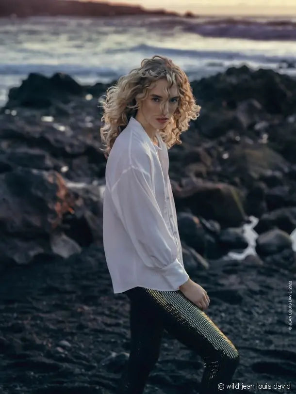 Uma mulher loira com cabelo encaracolado, vestindo uma camisa branca e calças escuras brilhantes, caminha numa praia rochosa ao pôr do sol.
