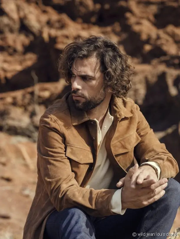 
Um homem com cabelo e barba castanhos e encaracolados, vestindo um casaco de camurça castanho, uma camisa clara e calças de ganga, sentado num cenário rochoso, olhando para o lado.