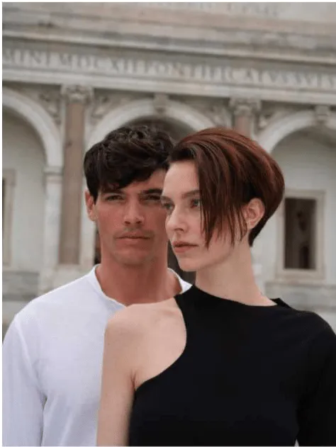 Um homem e uma mulher posam juntos. O homem, com cabelo escuro, veste uma camisola branca e está em segundo plano. A mulher, em primeiro plano, tem cabelo curto e castanho-avermelhado e veste uma camisola preta de um ombro só. Ambos olham para a direita da imagem, com um edifício clássico em segundo plano.