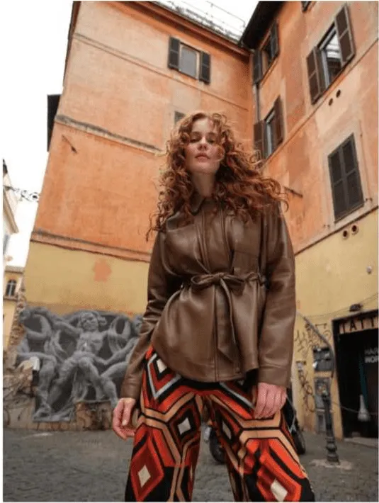 Uma mulher com cabelo ruivo e encaracolado, vestindo um casaco de couro castanho e calças com padrão geométrico em tons de laranja, vermelho e branco, posa na rua em frente a um edifício antigo de cor pêssego com um mural cinzento na parede.