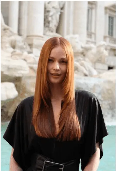 Uma mulher com cabelo ruivo longo e liso, vestindo uma blusa preta larga e um cinto preto, posa em frente à Fonte de Trevi. Ela olha diretamente para a câmara.