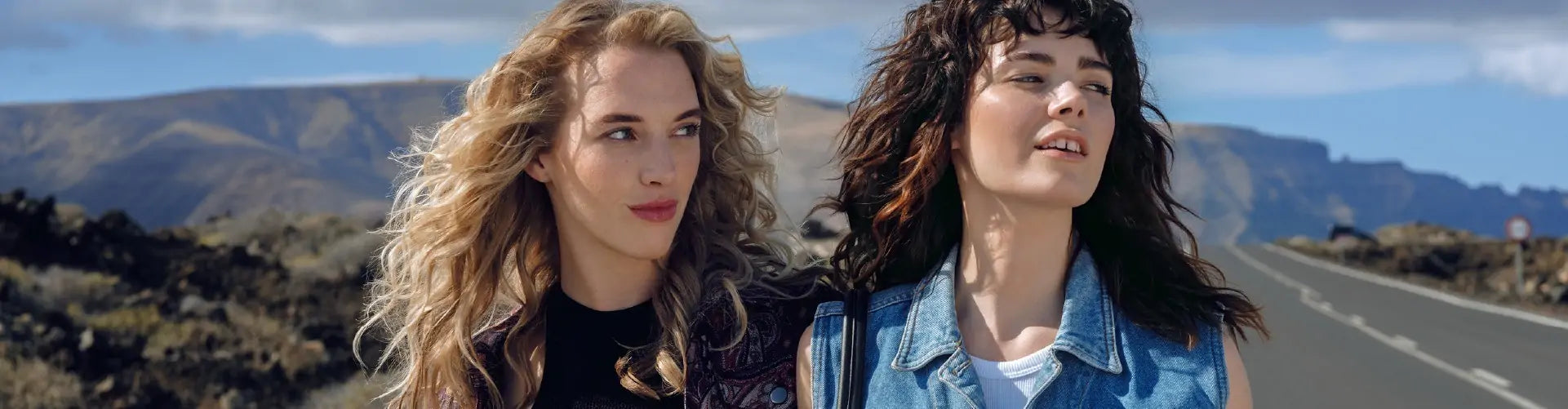 Duas mulheres jovens, uma loira com cabelo ondulado e outra morena com cabelo encaracolado, caminham ao longo de uma estrada com paisagens montanhosas ao fundo.