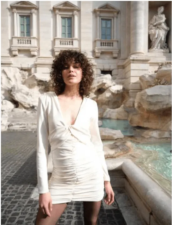 Uma mulher com cabelo castanho encaracolado, vestindo um vestido branco justo e meias pretas, posa em frente à Fonte de Trevi em Roma.