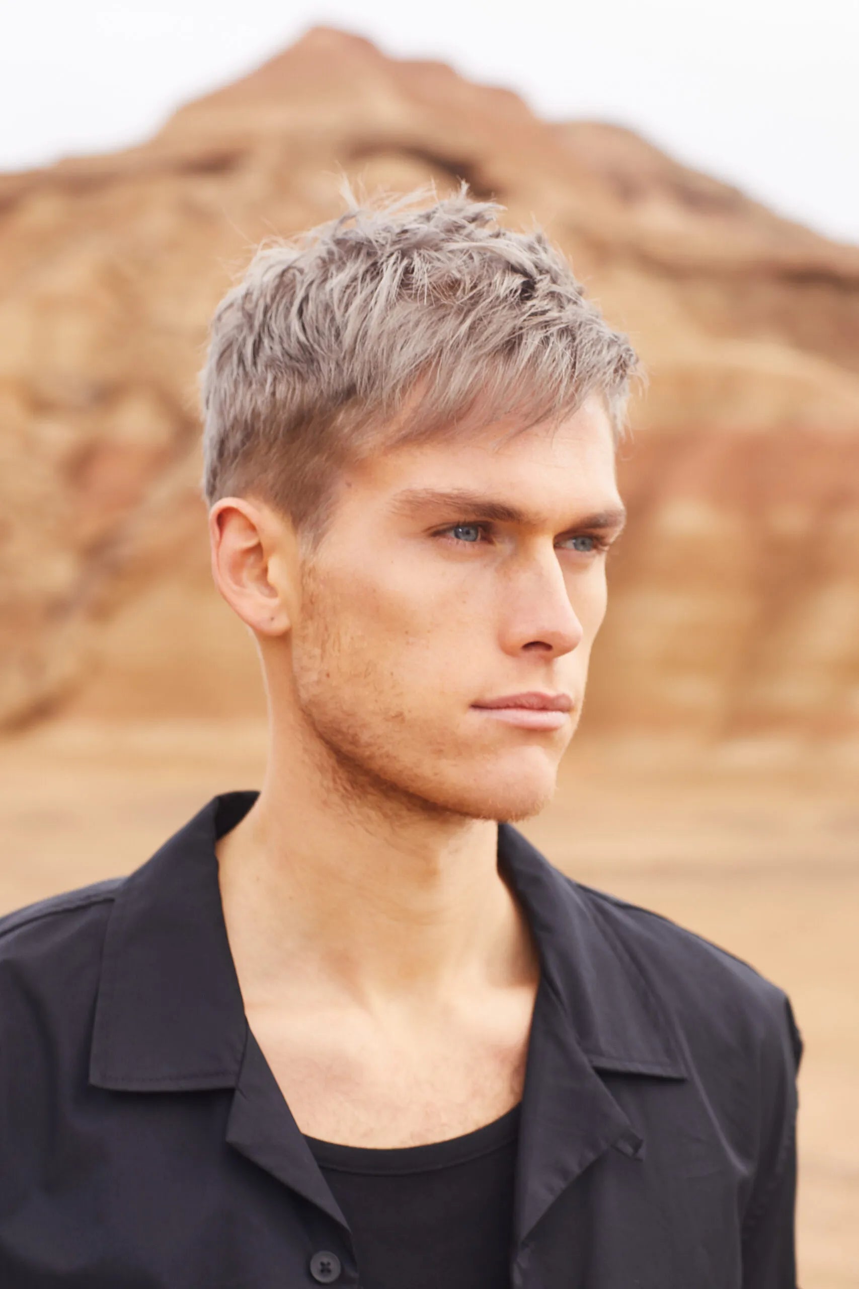 Um homem com cabelo loiro-platinado e olhos azuis, vestindo uma camisa preta, olha para o lado. O fundo é uma paisagem de montanhas arenosas.