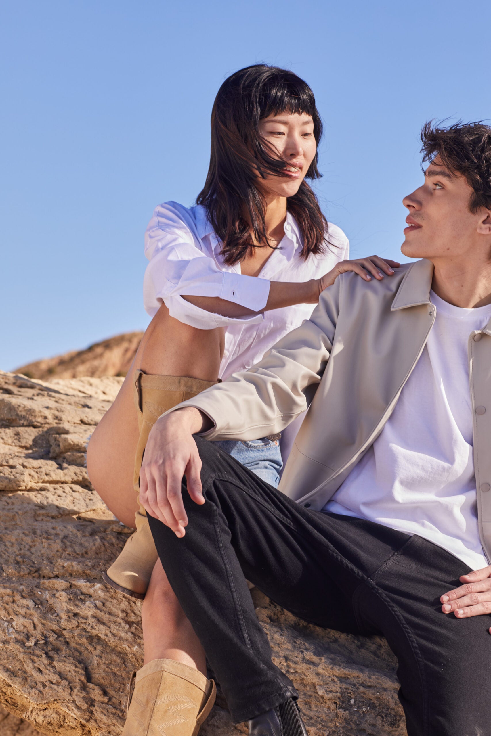 
Um homem e uma mulher posam juntos num cenário rochoso. A mulher, com cabelo escuro, veste uma camisa branca desabotoada e calções claros, e está de pé atrás do homem, com uma mão no ombro dele. O homem tem cabelo escuro, veste um casaco bege e calças pretas, e está sentado, olhando para a mulher.
