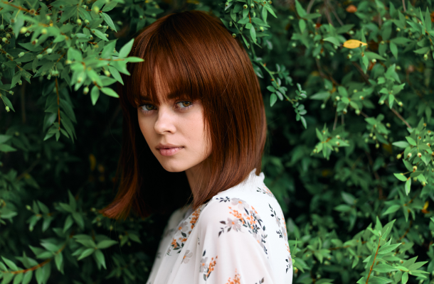 Mulher com cabelos ruivos, de pé entre uma exuberante folhagem verde, olha para longe